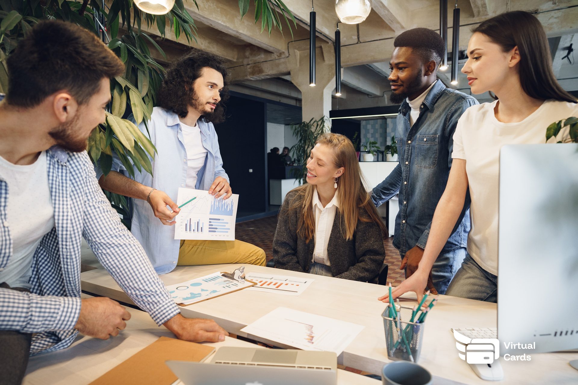 Five colleagues around a shared desk reviewing printed spending charts in an open-plan office, the kind of team that issues a separate card per commitment.